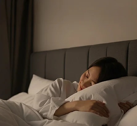 침대에 누워 있는 한 사람이 보이며, 베이지색 헤드보드가 있는 침실의 모습입니다. 사람은 흰색 잠옷을 입고 흰색 베개와 이불에 둘러싸여 있습니다. 방 안은 조용하고 편안한 분위기를 자아내며, 조명이 은은하게 비추고 있습니다.