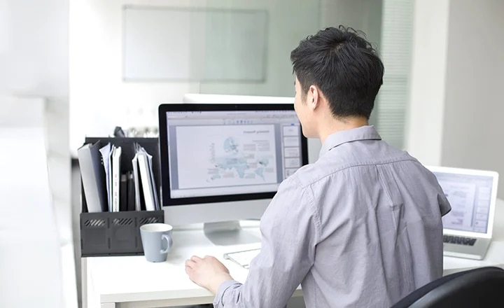 사무실 책상에 앉아 있는 남성이 컴퓨터 모니터를 보고 있습니다. 모니터에는 그래프와 텍스트가 포함된 화면이 표시되어 있으며, 책상 위에는 서류 정리함과 컵이 놓여 있습니다. 노트북도 함께 배치되어 있습니다.