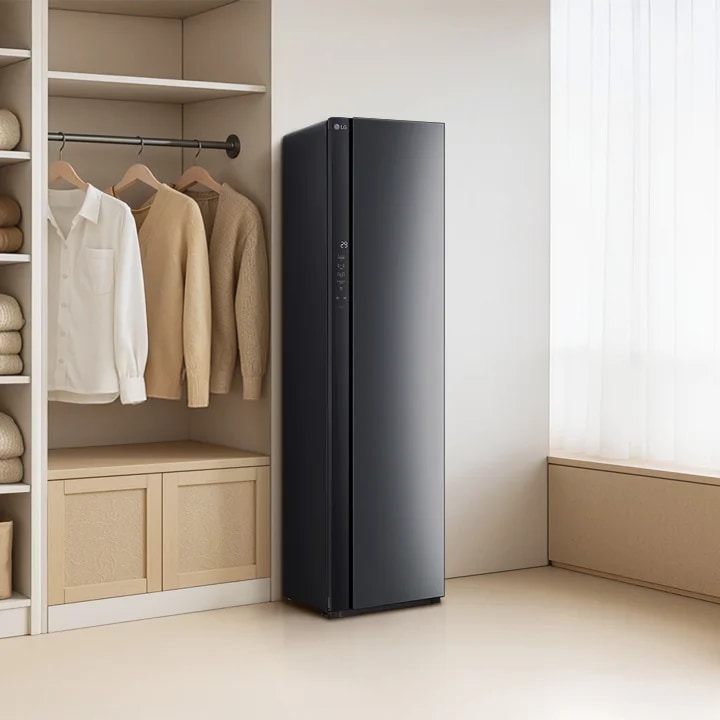 어두운 색상의 LG 스타일러가 설치되어 있으며, 그 옆에는 여러 벌의 옷이 걸려 있습니다. 스타일러 옆에는 나무로 된 서랍과 선반이 있으며, 그 위에는 가방과 상자가 놓여 있습니다. 오른쪽에는 커튼이 드리워져 있고, 왼쪽에는 베이지색 침대와 액자가 걸려 있는 벽이 보입니다. 침대 옆에는 책과 꽃병이 놓여 있습니다.